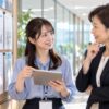 社内の廊下でタブレットの募集一覧を見ながら上司と話す女性社員